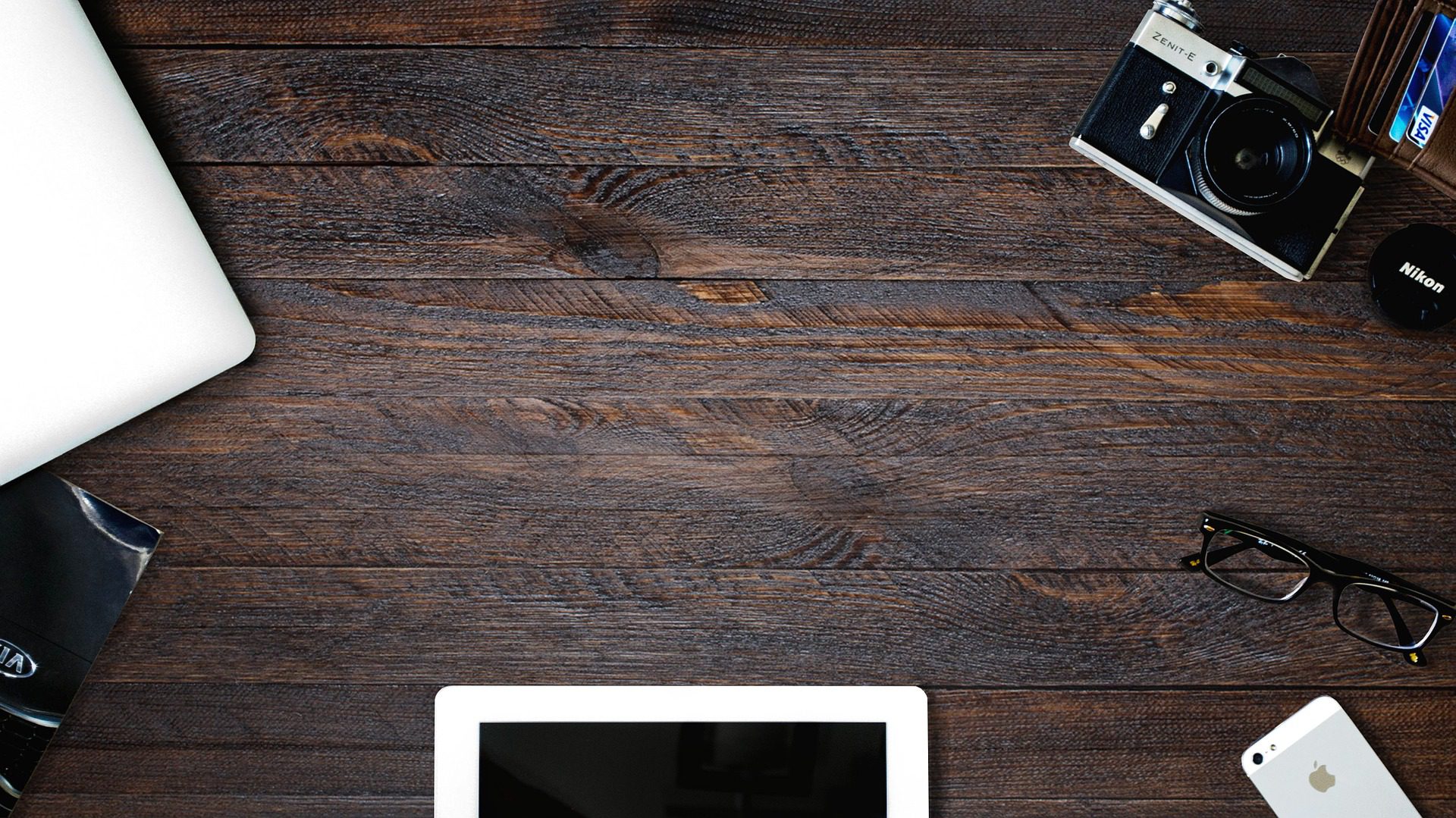 A desk with a tablet, laptop, camera, wallet, glasses, and iPhone