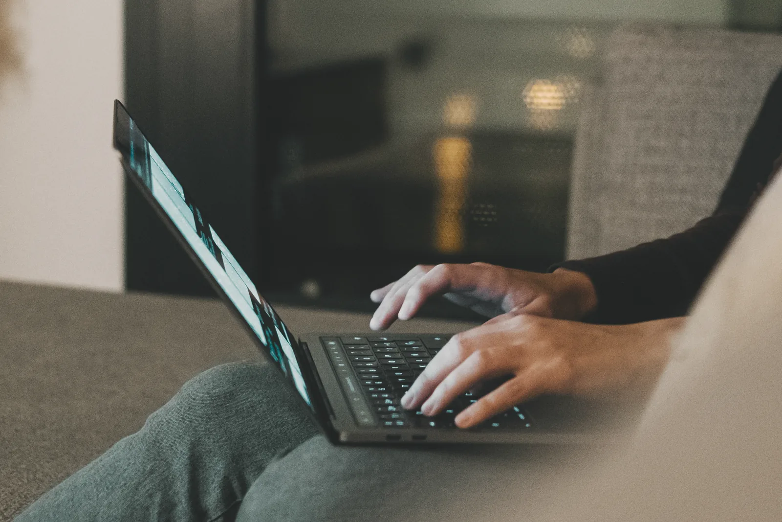 Person typing on a laptop keyboard while seated, the screen glowing in a dim room