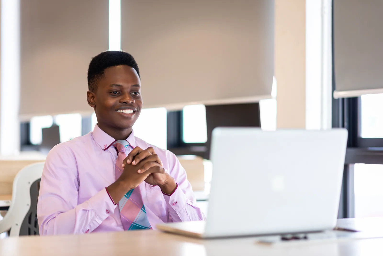 Smiling man in a pink shirt and patterned tie with hands clasped, looking at an open laptop in a bright office