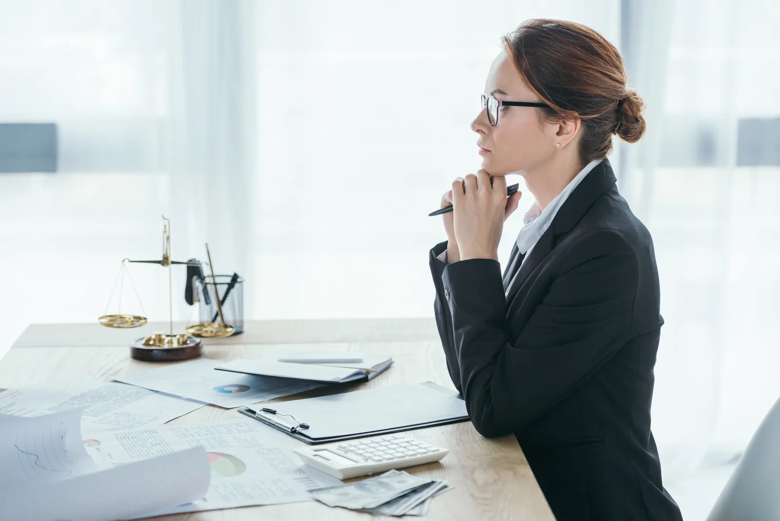 Lawyer in a dark suit resting her chin on her hand at a desk with brass scales of justice, charts, a calculator and cash